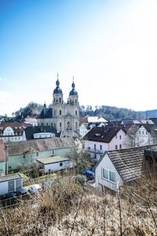 A quaint village scene depicting a series of residential houses with varying roof styles and colors. Dominating the backdrop is a grand, historic church with two tall towers, each topped with an ornate spire. The village is surrounded by rolling hills and forests, under a clear blue sky.