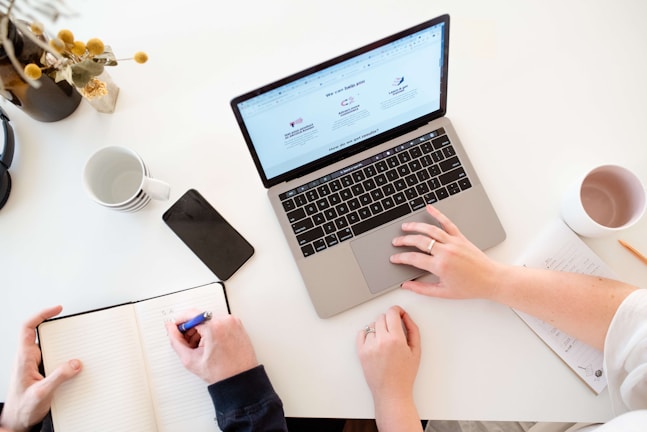 A workspace setting featuring a laptop displaying a webpage, alongside a notebook where one person is writing with a pen. Another person is using the laptop. Surrounding items include a smartphone, coffee mugs, a vase with flowers, and a piece of paper with notes. The scene is brightly lit with a minimalist aesthetic.