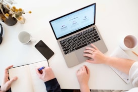 A workspace setting featuring a laptop displaying a webpage, alongside a notebook where one person is writing with a pen. Another person is using the laptop. Surrounding items include a smartphone, coffee mugs, a vase with flowers, and a piece of paper with notes. The scene is brightly lit with a minimalist aesthetic.