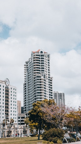 A tall, modern residential skyscraper with numerous balconies stands prominently against a partly cloudy sky. The building is surrounded by other urban structures and lush greenery, including a large tree in the foreground. The scene captures a mix of urban architecture and natural elements.