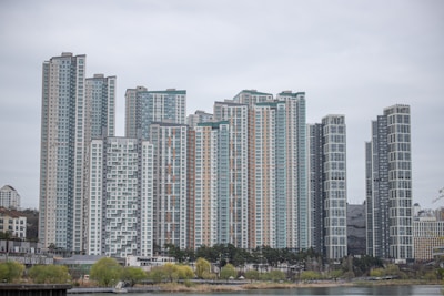 A collection of modern high-rise apartment buildings stands prominently in an urban setting. The skyscrapers have a variety of window patterns and are mostly white and grey, with some colored accents. In the foreground, trees with green foliage and a body of water provide contrast to the architectural lines.