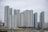 A collection of modern high-rise apartment buildings stands prominently in an urban setting. The skyscrapers have a variety of window patterns and are mostly white and grey, with some colored accents. In the foreground, trees with green foliage and a body of water provide contrast to the architectural lines.