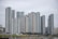 A collection of modern high-rise apartment buildings stands prominently in an urban setting. The skyscrapers have a variety of window patterns and are mostly white and grey, with some colored accents. In the foreground, trees with green foliage and a body of water provide contrast to the architectural lines.