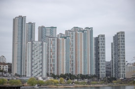 A collection of modern high-rise apartment buildings stands prominently in an urban setting. The skyscrapers have a variety of window patterns and are mostly white and grey, with some colored accents. In the foreground, trees with green foliage and a body of water provide contrast to the architectural lines.