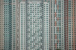 A large cluster of high-rise apartment buildings with varied window designs. They are tightly packed together, featuring a mixture of beige, teal, white, and orange color schemes. The buildings have a somewhat repetitive and geometric appearance.