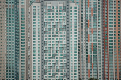 A large cluster of high-rise apartment buildings with varied window designs. They are tightly packed together, featuring a mixture of beige, teal, white, and orange color schemes. The buildings have a somewhat repetitive and geometric appearance.