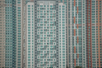 A large cluster of high-rise apartment buildings with varied window designs. They are tightly packed together, featuring a mixture of beige, teal, white, and orange color schemes. The buildings have a somewhat repetitive and geometric appearance.