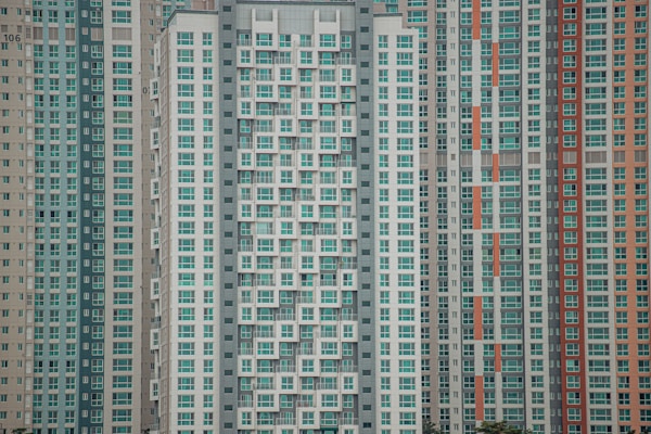 A large cluster of high-rise apartment buildings with varied window designs. They are tightly packed together, featuring a mixture of beige, teal, white, and orange color schemes. The buildings have a somewhat repetitive and geometric appearance.