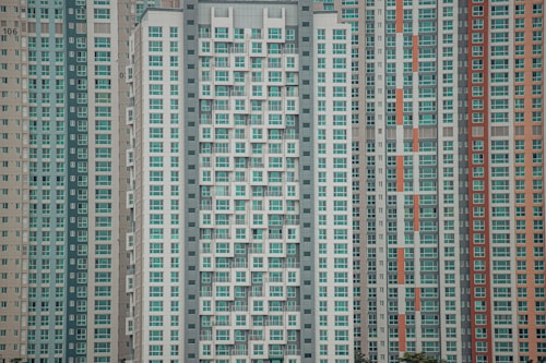 A large cluster of high-rise apartment buildings with varied window designs. They are tightly packed together, featuring a mixture of beige, teal, white, and orange color schemes. The buildings have a somewhat repetitive and geometric appearance.