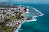An aerial view of a coastal area with a blend of urban development and natural landscapes. The coastline features white sandy beaches, bordered by clear turquoise and deep blue ocean waters. Numerous buildings are distributed throughout the landscape, indicating a residential or vacation resort area. Vegetation is prevalent, adding to the lush scenery. Distant mountains are visible on the horizon under a partly cloudy sky.