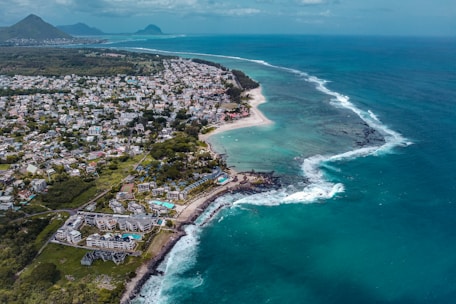 An aerial view of a coastal area with a blend of urban development and natural landscapes. The coastline features white sandy beaches, bordered by clear turquoise and deep blue ocean waters. Numerous buildings are distributed throughout the landscape, indicating a residential or vacation resort area. Vegetation is prevalent, adding to the lush scenery. Distant mountains are visible on the horizon under a partly cloudy sky.