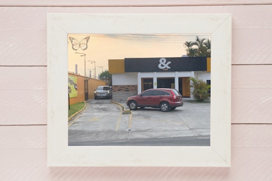 A framed photo features a caf&eacute; with a parking lot in front. A red SUV is parked in the center, and another vehicle is at the side near an orange wall. The building has a modern design with a black and yellow color scheme and a logo that appears to be an ampersand above the word 'CAFE'. There is a decorative butterfly in the top left corner of the frame.
