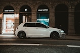 A white car is parked on an urban street in front of high-end luxury store windows displaying 'Louis Vuitton.' The shopfronts are dimly lit, and there are elegant architectural details above. The scene is captured at night, giving a sophisticated and sleek atmosphere.