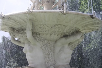 A decorative fountain featuring a classical stone sculpture of figures supporting a large basin. Water cascades down from the basin, creating a dynamic and cooling effect. The scene includes lush greenery in the background, illuminated by sunlight, enhancing the serene atmosphere.