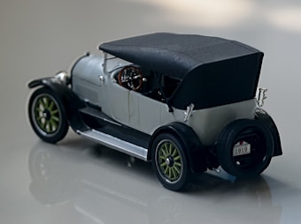 A vintage model car featuring an early 20th-century design with a soft black top. The vehicle has green spoked wheels and a visible spare tire at the rear. A steering wheel and instrumental panel are visible inside, with a license plate showing the year 1919.