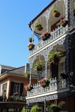 An elegant wrought iron balcony with hanging flower baskets is prominently featured on a building with classic architectural elements. The bright blue sky contrasts with the vibrant green plants and colorful flowers, creating a lively and inviting atmosphere.