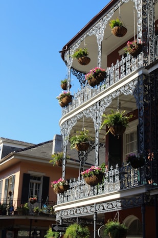 An elegant wrought iron balcony with hanging flower baskets is prominently featured on a building with classic architectural elements. The bright blue sky contrasts with the vibrant green plants and colorful flowers, creating a lively and inviting atmosphere.