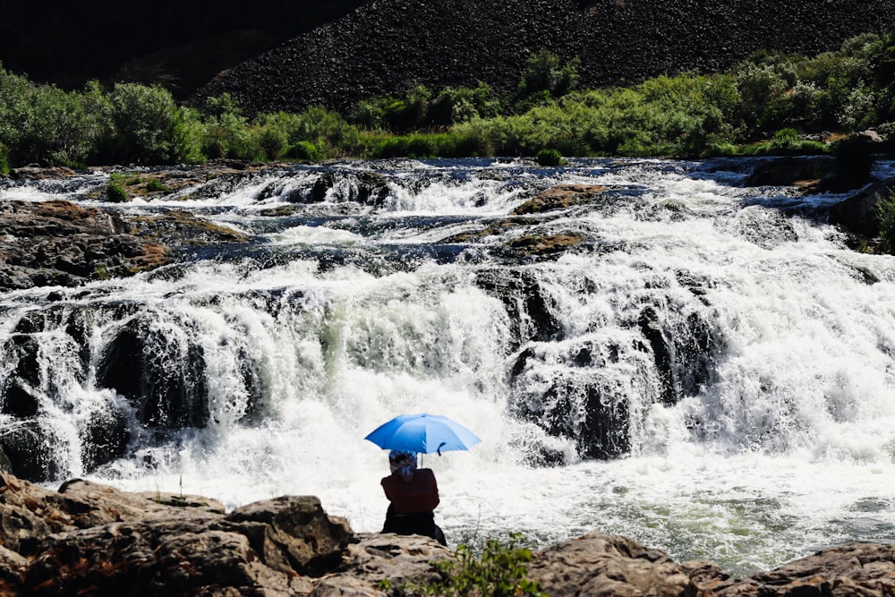 a person sitting on a rock with an umbrella in front of a waterfall