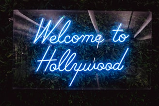 A vibrant neon sign displays the words 'Welcome to Hollywood' in bright blue light, set against a backdrop of lush green foliage, creating a striking contrast that draws attention.