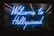 A vibrant neon sign displays the words 'Welcome to Hollywood' in bright blue light, set against a backdrop of lush green foliage, creating a striking contrast that draws attention.