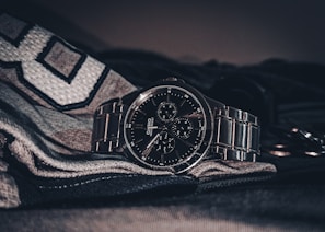 A sleek, metallic wristwatch with a dark face and multiple dials is placed on a textured fabric surface. The watch features a polished metal strap, and the surrounding environment is slightly dim, highlighting the watch's reflective surfaces.