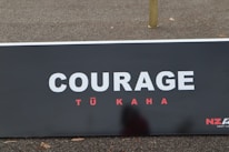 A large signboard displays the word 'COURAGE' in bold white letters with the words 'TŪ KAHA' in smaller red letters underneath, set against a dark background. The ground is visible with some scattered leaves.