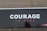 A large signboard displays the word 'COURAGE' in bold white letters with the words 'TŪ KAHA' in smaller red letters underneath, set against a dark background. The ground is visible with some scattered leaves.