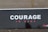 A large signboard displays the word 'COURAGE' in bold white letters with the words 'TŪ KAHA' in smaller red letters underneath, set against a dark background. The ground is visible with some scattered leaves.