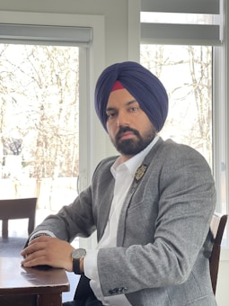 A man wearing a gray blazer and a turban sits at a wooden table in a room with large windows. The background shows leafless trees, indicating a possible winter or early spring setting. He appears to be looking calmly at the camera, wearing a watch on his right wrist.