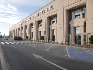 A large, modern airport building with a prominent sign reading 'Aeropuerto de Malaga Costa del Sol'. The structure has multiple windows, pillars, and an entrance area, with flags on the facade. Vehicles including cars are present on the adjacent road, along with a pedestrian crossing and parking spaces.