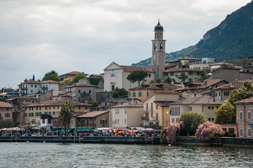 Charming lakeside village with colorful buildings and a prominent church tower, surrounded by lush greenery and mountains in the background. The calm water reflects the serene, picturesque landscape. Flags line the waterfront, adding vibrant pops of color to the scene.