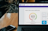 A laptop displays a credit score website with a score of 825, labeled as excellent, set on a wooden table with a smartphone nearby. Dark leafy plant elements are blurred in the background.