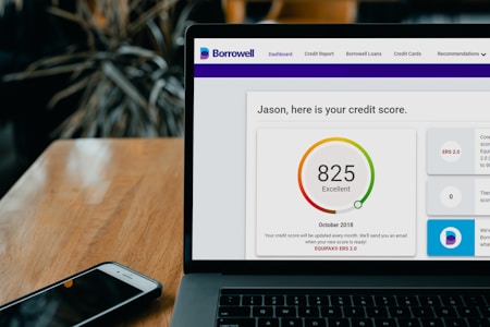 A laptop displays a credit score website with a score of 825, labeled as excellent, set on a wooden table with a smartphone nearby. Dark leafy plant elements are blurred in the background.