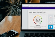 A laptop displays a credit score website with a score of 825, labeled as excellent, set on a wooden table with a smartphone nearby. Dark leafy plant elements are blurred in the background.