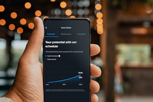 A hand is holding a smartphone displaying a financial application. The screen shows a chart with a growth prediction in percentage, and sections labeled Personal, Retirement, and Custodial. The chart predicts cash invested and saved over a period of time, with specific dollar values provided for future growth.