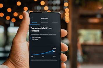 A hand is holding a smartphone displaying a financial application. The screen shows a chart with a growth prediction in percentage, and sections labeled Personal, Retirement, and Custodial. The chart predicts cash invested and saved over a period of time, with specific dollar values provided for future growth.