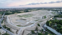 An aerial view of a large, partially constructed or barren area surrounded by a road with adjoining small green patches and trees. In the background, there are residential areas and a stretch of greenery extending to the horizon. The sky is lightly clouded with patches of both blue and orange tones, suggesting a sunset or sunrise.