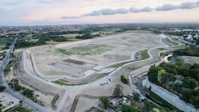 An aerial view of a large, partially constructed or barren area surrounded by a road with adjoining small green patches and trees. In the background, there are residential areas and a stretch of greenery extending to the horizon. The sky is lightly clouded with patches of both blue and orange tones, suggesting a sunset or sunrise.