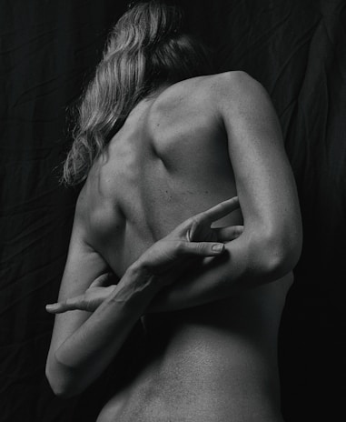A monochrome photograph capturing the back of a person with long hair. The individual is standing against a dark background, and their arms are bent in an artistic pose. The lighting enhances the contours and textures of the skin.