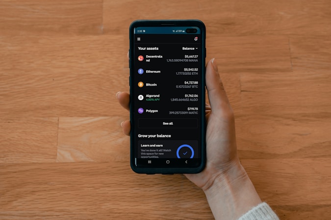 A hand holds a smartphone displaying a cryptocurrency portfolio with various assets including Decentraland, Ethereum, Bitcoin, Algorand, and Polygon. The balance for each asset is shown alongside icons representing each cryptocurrency.