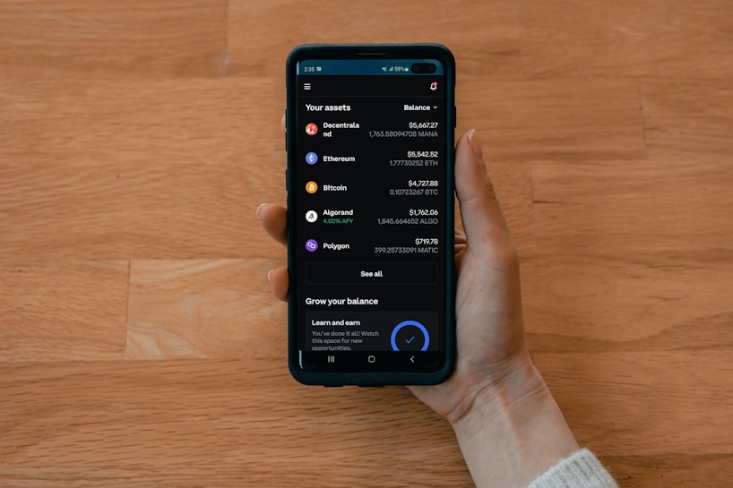 A hand holds a smartphone displaying a cryptocurrency portfolio with various assets including Decentraland, Ethereum, Bitcoin, Algorand, and Polygon. The balance for each asset is shown alongside icons representing each cryptocurrency.