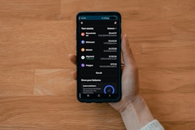 A hand holds a smartphone displaying a cryptocurrency portfolio with various assets including Decentraland, Ethereum, Bitcoin, Algorand, and Polygon. The balance for each asset is shown alongside icons representing each cryptocurrency.