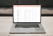 A laptop on a table displaying a cryptocurrency trading platform. The screen lists various cryptocurrencies along with their buy and sell values, spread percentages, and 24-hour change percentages. The keyboard and touchpad of the laptop are visible, set against a neutral background with a sofa in the back.