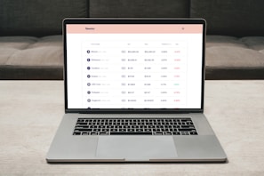 A laptop on a table displaying a cryptocurrency trading platform. The screen lists various cryptocurrencies along with their buy and sell values, spread percentages, and 24-hour change percentages. The keyboard and touchpad of the laptop are visible, set against a neutral background with a sofa in the back.