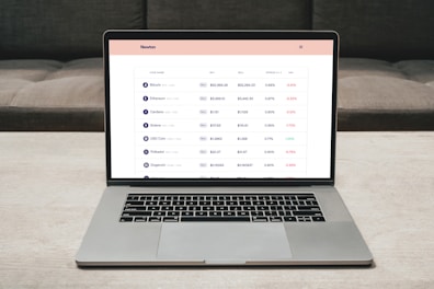 A laptop on a table displaying a cryptocurrency trading platform. The screen lists various cryptocurrencies along with their buy and sell values, spread percentages, and 24-hour change percentages. The keyboard and touchpad of the laptop are visible, set against a neutral background with a sofa in the back.