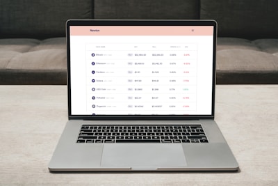 A laptop on a table displaying a cryptocurrency trading platform. The screen lists various cryptocurrencies along with their buy and sell values, spread percentages, and 24-hour change percentages. The keyboard and touchpad of the laptop are visible, set against a neutral background with a sofa in the back.