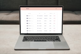 A laptop on a table displaying a cryptocurrency trading platform. The screen lists various cryptocurrencies along with their buy and sell values, spread percentages, and 24-hour change percentages. The keyboard and touchpad of the laptop are visible, set against a neutral background with a sofa in the back.