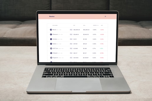 A laptop on a table displaying a cryptocurrency trading platform. The screen lists various cryptocurrencies along with their buy and sell values, spread percentages, and 24-hour change percentages. The keyboard and touchpad of the laptop are visible, set against a neutral background with a sofa in the back.