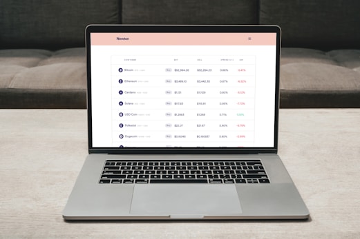 A laptop on a table displaying a cryptocurrency trading platform. The screen lists various cryptocurrencies along with their buy and sell values, spread percentages, and 24-hour change percentages. The keyboard and touchpad of the laptop are visible, set against a neutral background with a sofa in the back.