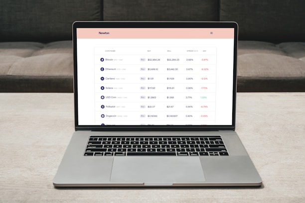 A laptop on a table displaying a cryptocurrency trading platform. The screen lists various cryptocurrencies along with their buy and sell values, spread percentages, and 24-hour change percentages. The keyboard and touchpad of the laptop are visible, set against a neutral background with a sofa in the back.